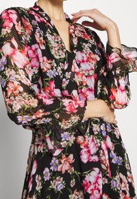 Czarna sukienka w kwiaty z długimi przezroczystymi rękawami, ozdobiona różowymi i niebieskimi kwiatami. Krój sukienki podkreśla talię i ma dekolt w kształcie litery V.