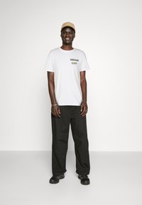 Camiseta blanca con el texto "RIDE HARD", combinada con pantalones sueltos negros, gorra beige y zapatillas negras sobre un fondo neutro.