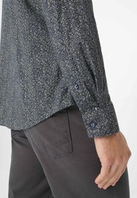 Mann in einem dunkelblumigen Langarmhemd mit geknöpften Manschetten und dunkelgrauen Hosen, die Hand entspannt an der Seite.