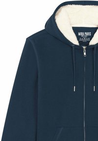 Hoodie zippé bleu marine avec une capuche douce en sherpa crème, des poches avant et des matériels en argent pour la fermeture éclair et les cordons.