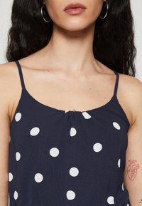 Top sans manches bleu marine avec des pois blancs, présentant des fines bretelles et un col froncé. Texture de tissu lisse et léger.