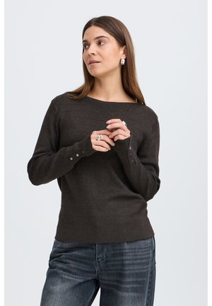Vrouw met lang bruin haar, draagt een donkere trui met knoopdetails en een spijkerbroek, staat met de handen gevouwen en kijkt naar links.