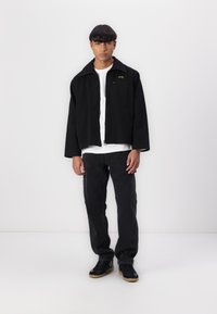 Svart jakke med spiss krage og glidelås, brukt over en hvit skjorte. Kombinert med svarte jeans og svarte sneakers med gummisåler.