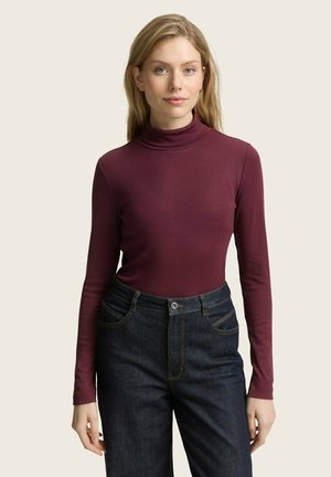 Nainen, joka on pukeutunut tiukkaan burgundinpunaisiin turtleneck-paitaan, jossa on pitkät hihat, ja hänellä on päällään korkeavyötäröiset tummansiniset farkut yksinkertaista taustaa vasten.