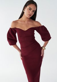 Vīnsarkanā kleita bez pleciem ar uzpūstām piedurknēm, pieguļošu korseti un teksturētu audumu. Zelta aproce uz plaukstas.