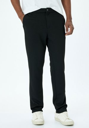 Pantalons stretch noirs avec une texture lisse, design slim fit, poches avant et fermeture à bouton, accompagnés de baskets blanches.
