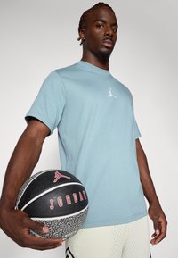 Gaiši zils īsais piedurknes T-krekls ar baltu Jordan logo. Individuāls tur melnu basketbola ar sarkaniem un baltajiem akcentiem. Tekstūrēta adīta auduma.