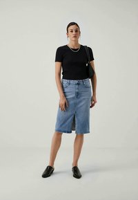 Sieviete, kas ģērbusies melnā īsās piedurknes topā, gaiši zilā ceļgala garuma džinsu svārkos ar priekšējo izgriezumu, melnās slīdšus kurpes un pleca somu, stāv neitrālā pozā.