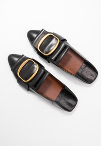 Chaussures plates en cuir noir à bout carré, dotées de grandes boucles dorées et de semelles intérieures marron, disposées côte à côte sur un fond blanc.