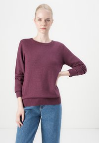 Merinoulltröja i mörk mauve, rund halsringning, ribbade ärmslut och nederkant, kombinerad med ljusblå jeans, uppställd på en neutral bakgrund.