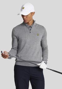 Grauer Pullover mit langen Ärmeln und Reißverschlusskragen sowie gelbem Logo, kombiniert mit einem Hemd mit Kragen. Eine weiße Baseballmütze und ein Golfschläger in der Hand.