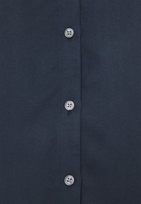 Mørkeblå skjorte med tekstureret stof. Har en række af fire sølvknapper med et fire-huls design langs placket.