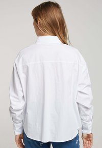 Chemise blanche à manches longues avec un col, couture de empiècement au dos et un ourlet arrondi. Le tissu semble lisse et légèrement texturé.
