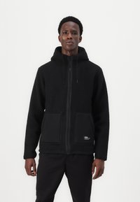 Veste en polaire noire avec fermeture éclair à l'avant, capuche ajustable et grandes poches avant. Tissu texturé avec un panneau noir lisse aux niveaux des poches.