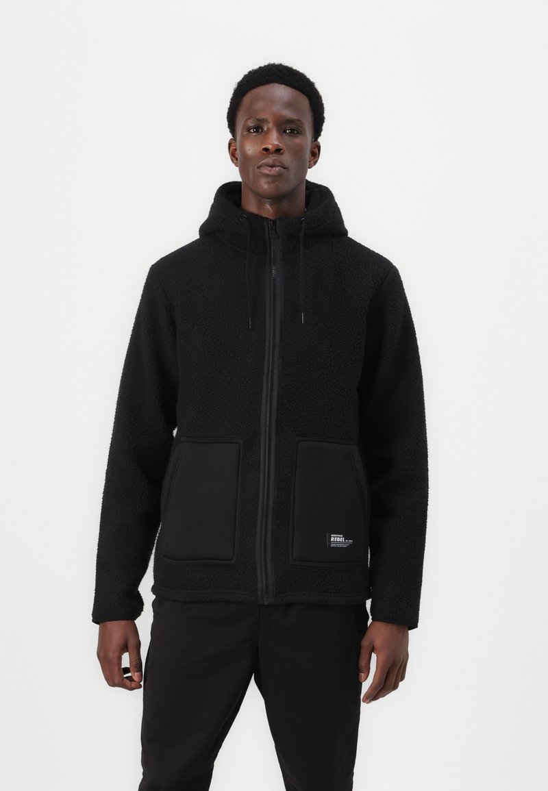 Veste en polaire noire avec fermeture éclair à l'avant, capuche ajustable et grandes poches avant. Tissu texturé avec un panneau noir lisse aux niveaux des poches.