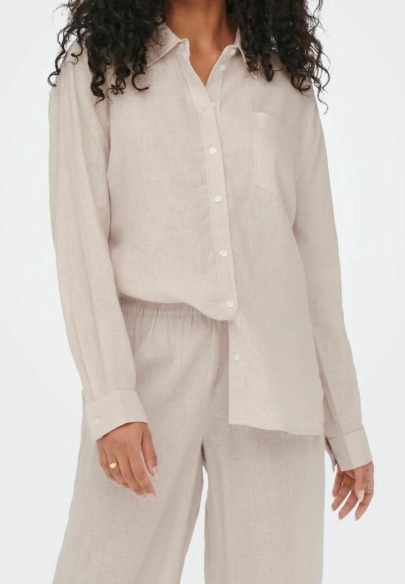 Camisa de lino beige claro con mangas largas, cierre de botones y un bolsillo frontal. Corte relajado, combinada con pantalones de lino sueltos a juego.
