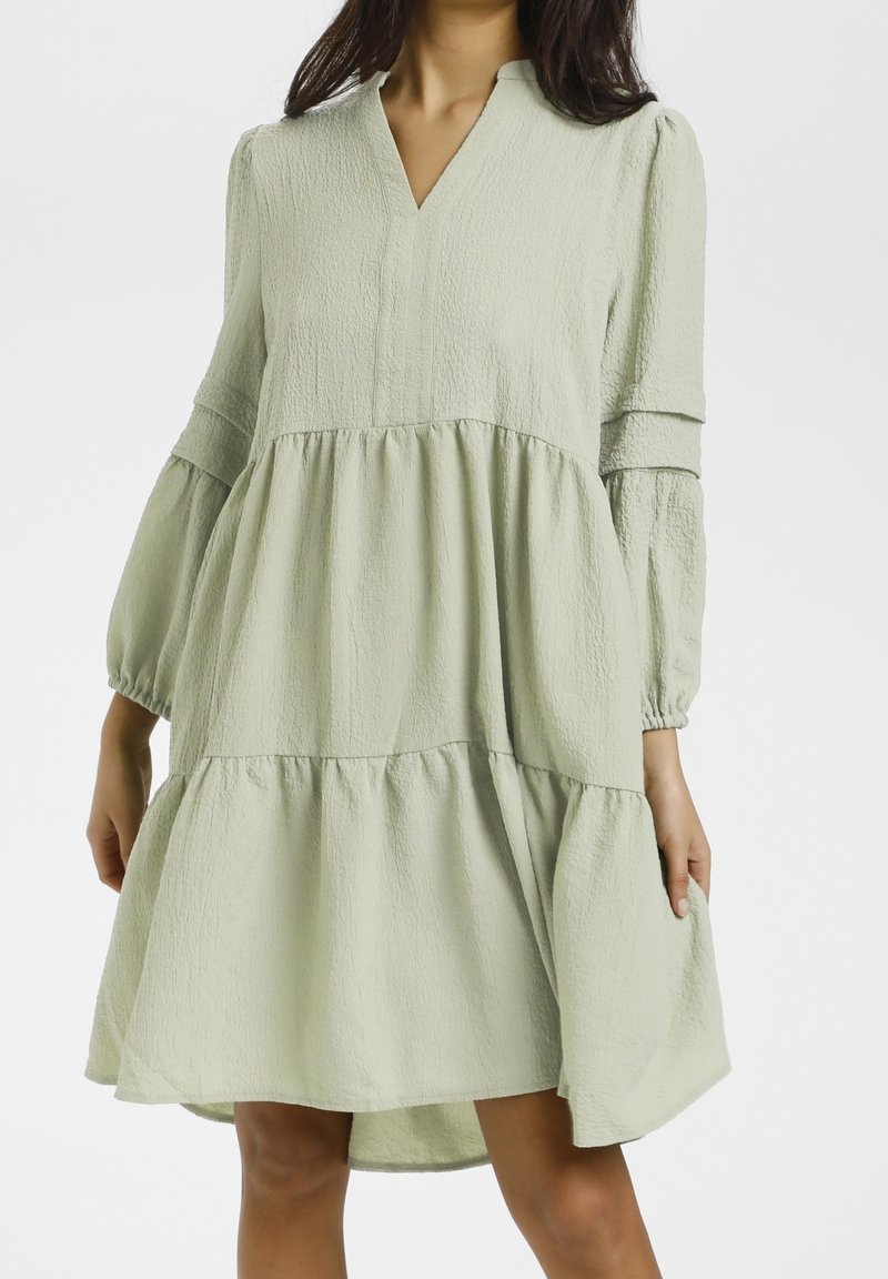 Robe vert clair texturée avec un col en V et un ourlet à volants. Présente des manches longues bouffantes et une coupe décontractée. Matière douce et fluide.