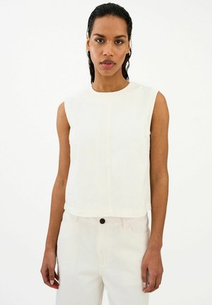 Femme aux cheveux mouillés portant un haut blanc sans manches et un pantalon blanc taille haute assorti, sur un fond blanc uni.