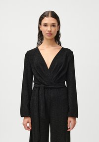 Czarny kombinezon z long sleeves, głębokim dekoltem w kształcie V, fasonem kopertowym i paskiem w talii. Materiał ma błyszczącą teksturę, która odbija światło.