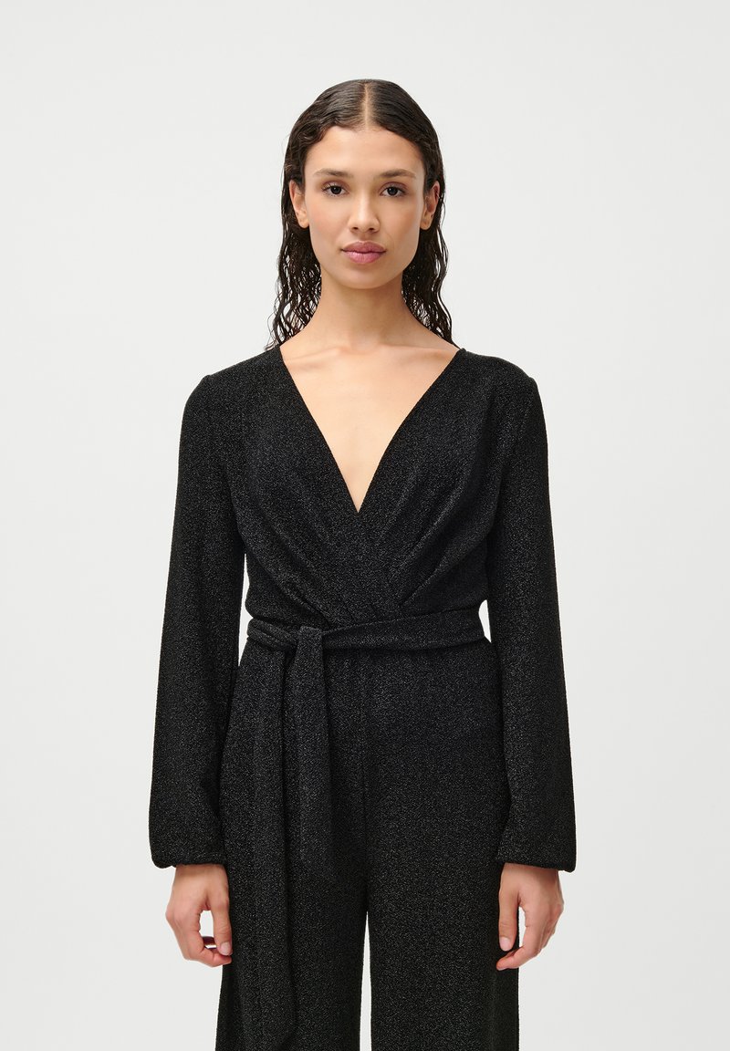 Crni kombinezon s dugim rukavima, dubokim V-izrezom, omotani dizajn i pojasom u struku. Tkanina ima sjajnu teksturu koja odražava svjetlost.
