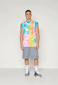 Színes ujjatlan ing batikmintával, rózsaszín, sárga és kék színekben, szürke rövidnadrággal és fekete sportcipővel párosítva.