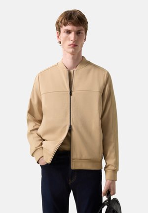 Junger Mann trägt eine beigefarbene Jacke mit Reißverschluss und dunkle Jeans, hält einen schwarzen Rucksack am Griff mit neutralem Gesichtsausdruck.