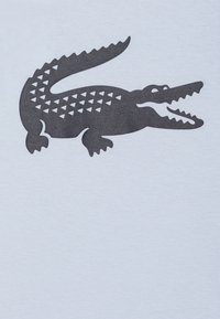 T-shirt bleu clair avec un graphique de crocodile gris foncé, défini par des motifs triangulaires et des détails marquants, y compris des mâchoires ouvertes et une queue.