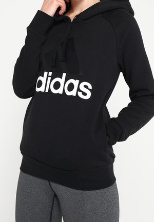 Personne portant un sweat à capuche Adidas noir avec un logo blanc et des leggings gris, une main touchant le visage et l'autre dans la poche du sweat.