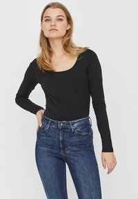 Bodysuit noir à manches longues avec un col rond, associé à un jean en denim bleu à la silhouette ajustée et présentant des détails de surpiqûres visibles.