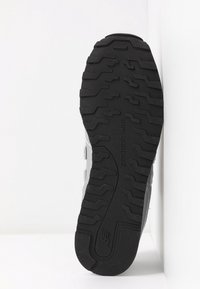 Semelle en caoutchouc noire d’une basket avec des rainures profondes en zigzag, logo New Balance embossé près du centre, chaussure sur fond blanc.