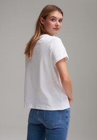 Witte katoenen t-shirt met opgerolde korte mouwen, recht gesneden en een ronde hals. Te dragen met blauwe denim jeans, getoond vanuit een lichte achteraanzicht.