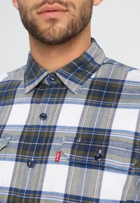 Camisa de franela a cuadros en colores azul, verde y blanco, con cuello abotonado y dos bolsillos en el pecho. Incluye detalle de etiqueta de marca.