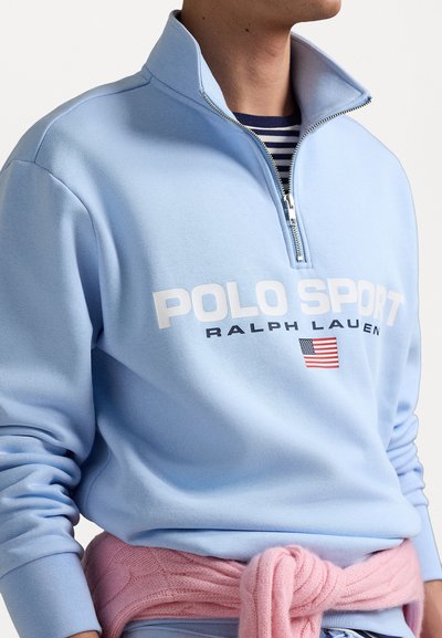 Jasnoniebieski sweter z ćwierć zamkiem z białym napisem "POLO SPORT", charakteryzujący się miękką teksturą i ściągaczami przy mankietach. Różowy sweter dzianinowy związany w pasie.