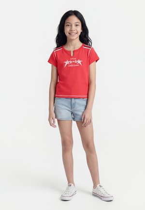 Egy lány áll, mosolyog, vörös rövid ujjú felsőt visel csillagos mintával, világos farmer rövidnadrágot és fehér tornacipőt fehér háttér előtt.