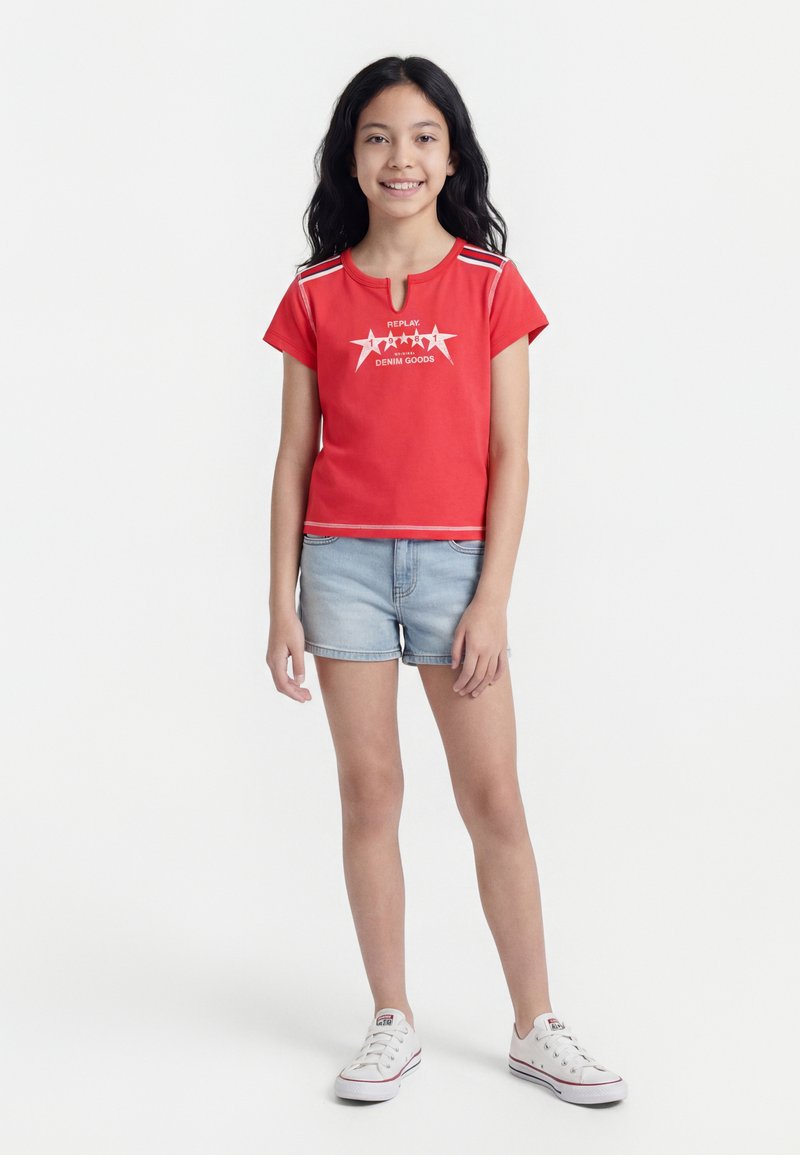 Κορίτσι που στέκεται και χαμογελά, φορώντας κόκκινο κοντομάνικο τοπ με γραφικά αστεριών, ανοιχτόχρωμα τζιν σορτς και λευκά αθλητικά, σε λευκό φόντο.