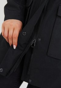 Chaqueta negra con cremallera hecha de tela suave con botones de presión, que cuenta con un cierre con cremallera y bolsillos laterales. Acentuada con un acabado texturizado.