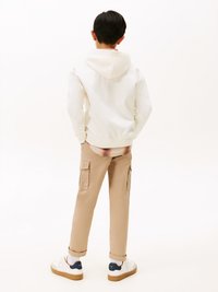 Gutt som står med hendene i lommene iført hvit hettegenser, beige cargo-bukser, hvite sneakers og en rutete skjorte delvis synlig under.