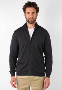 Sweatshirt zippé gris foncé avec un col montant, des poignets côtelés et une subtile touche de bleu le long de la fermeture éclair et de l'ourlet. Porté sur une chemise blanche.