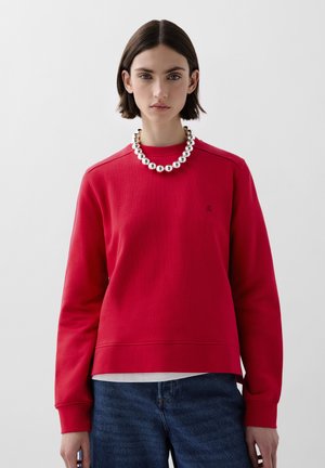 Sweatshirt rouge en coton avec texture côtelée, épaules tombantes et col rond. Associé à un collier en perles argentées et un jean en denim bleu.