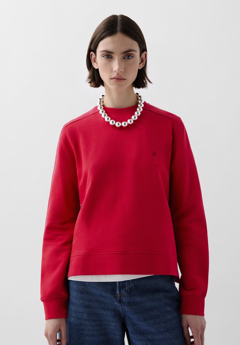 Rød bomulds-sweatshirt med ribbet tekstur, dropped skuldre og rund hals. Parret med en sølv perlekæde og blå denim jeans.