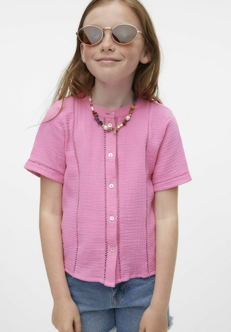 Camisa rosa de manga corta con cierre de botones, con tejido texturizado, costura decorativa y un corte relajado. Se lleva con pantalones cortos de mezclilla.