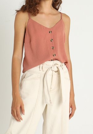 Femme portant un haut sans manches couleur rouille avec des boutons, associé à un pantalon taille haute à jambes larges de couleur blanc cassé, noué avec une ceinture en tissu.