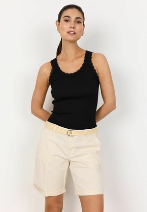 Woman wearing a black lace-trimmed sleeveless top and beige belted knee-length shorts, standing with hands behind her back.