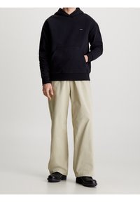 Sudadera negra con capucha y bolsillo frontal, combinada con pantalones de pierna ancha beige y calzado negro, todo sobre un fondo neutro.