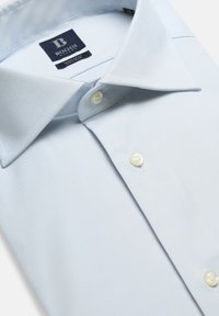 Ljusblå skjorta med en slät textur, spetsig krage och två vita knappar. Har en synlig varumärkesetikett inuti kragområdet.