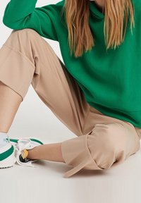 Personne assise, jambes croisées, portant un pantalon beige court, des baskets blanches avec des accents verts et un haut vert à manches longues.
