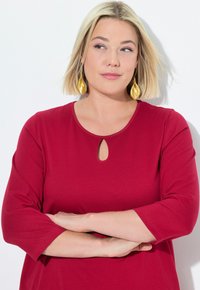 Punainen ribbipaita, jossa on kolmen neljänneksen hihat ja avaus kaula-aukossa. Mallilla on vaaleat hiukset ja kultaketjut korvissa.