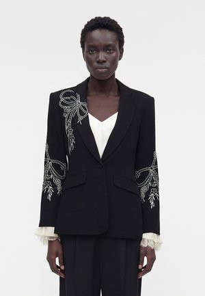 Γυναίκα φοράει μαύρο σακάκι με λευκή κεντημένη λουλουδάτη φιόγκο στους ώμους και τα μανίκια, φορεμένο πάνω από λευκό πουκάμισο με δαντελένια μανίκια.