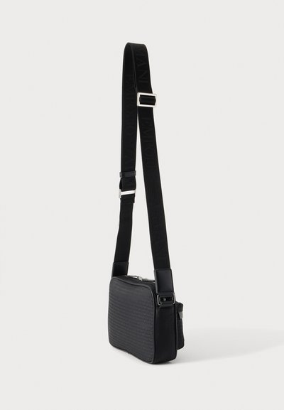 Μαύρη ορθογώνια τσάντα crossbody με ρυθμιζόμενο ιμάντα και διακριτικό ανάγλυφο λογότυπο στην υφασμάτινη επιφάνεια.