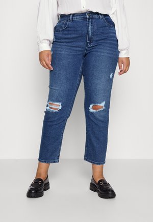 Person wearing blue ripped jeans, white long-sleeve blouse, and black loafers standing against a plain background.